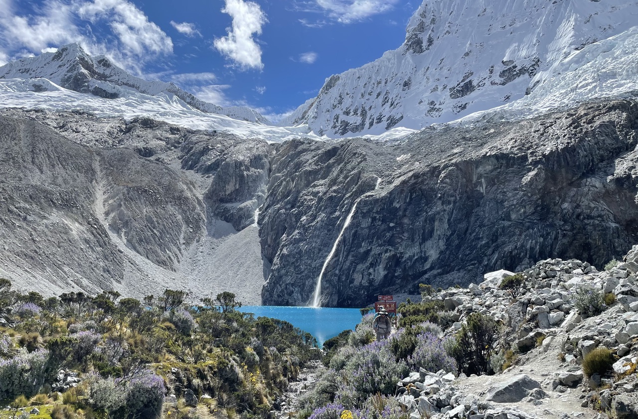 A turquoise glacial lake in a high Andean valley, ringed by snow-capped peaks and a thin waterfall, with scrubby alpine brush in the foreground