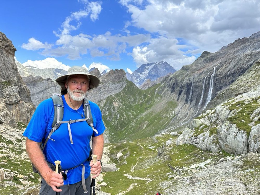 Jeffrey C. Hood backpacking on a mountain trail, with alpine peaks and a waterfall behind him